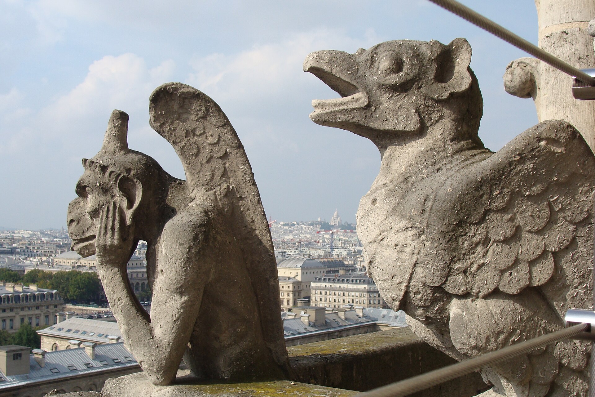 Two chimeras on the gallery between the west towers of Notre-Dame de Paris, with the dome of Sacré-Cœur visible on the skyline behind.