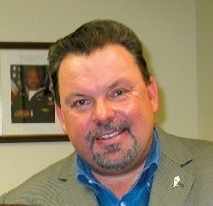 Head-and-shoulders photograph of Thomas Kinkade, a bearded man in a grey jacket over a blue shirt, smiling at the camera in an office with a framed photo behind him.